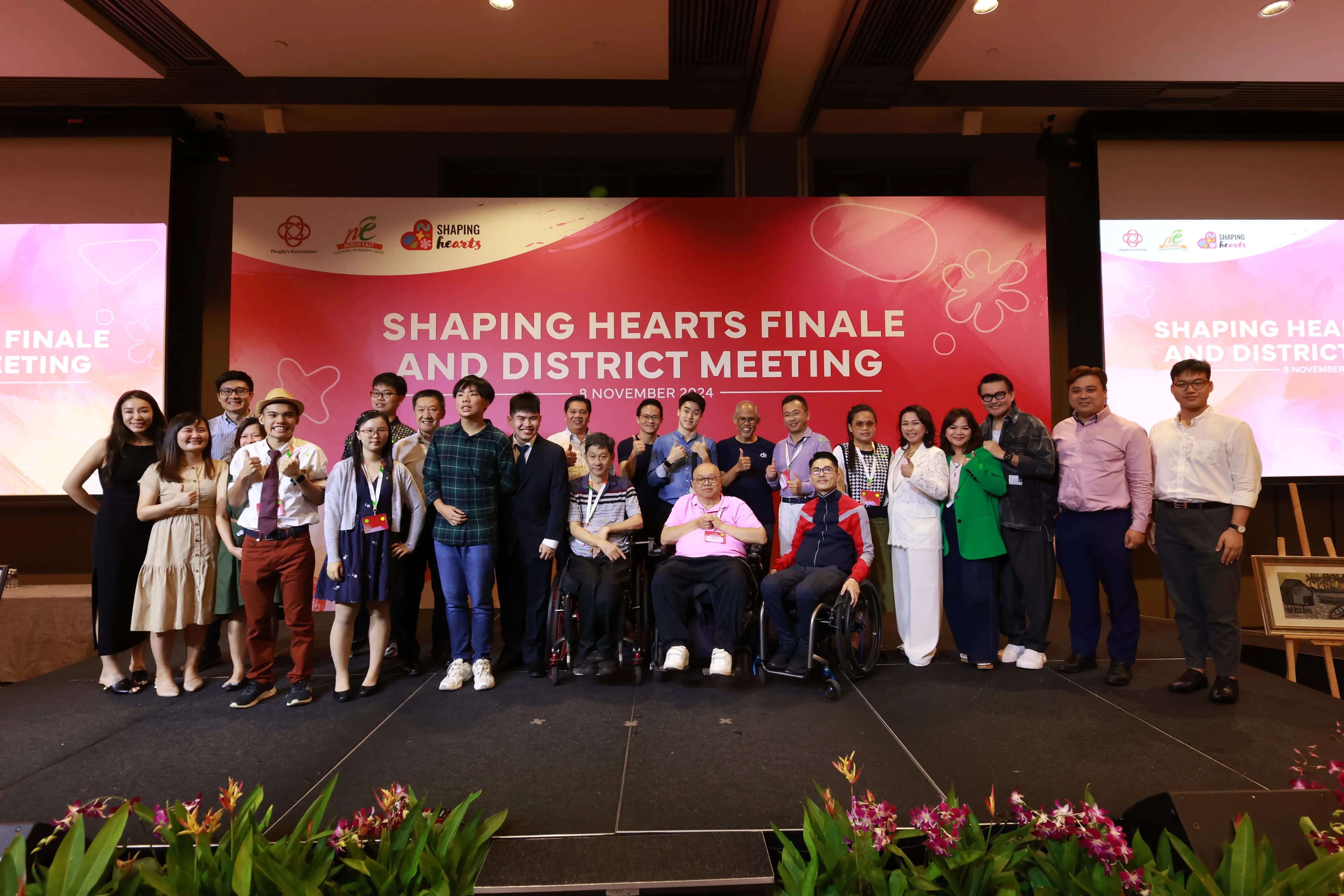 Group of people standing and sitting in front of a "Shaping Hearts Finale and District Meeting" banner on a stage.
